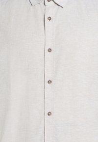 Ανοιχτό μπεζ πουκάμισο με κουμπιά, κλασικό γιακά και ορατά καφέ κουμπιά κατά μήκος του μπροστινού κέντρου.