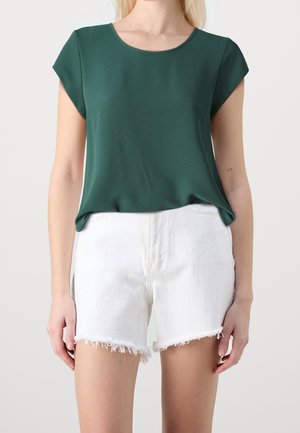 Kobieta ubrana w zieloną leśną bluzkę z krótkim rękawem włożoną do białych, postrzępionych szortów dżinsowych, stojąca na jednolitym tle.