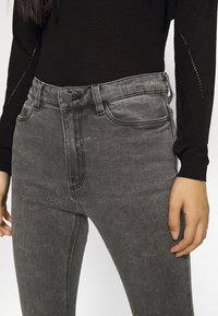 Jeans de mezclilla gris con corte ajustado, talle alto, diseño de cinco bolsillos, costuras visibles y cierre de botón en la parte delantera.