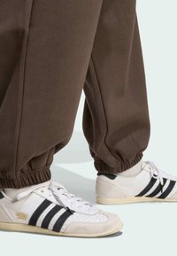 Bijele Adidas Japan tenisice s crnim prugama, nosene s smešastim hlačama na gumicu i bijelim čarapama na svijetloj pozadini.