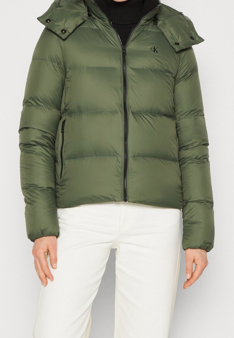 Kurtka typu puchowa w oliwkowym zielonym kolorze, zapinana na suwak z przodu, z odpinanym kapturem, kieszeniami na suwak oraz logo Calvin Klein na piersi. Gładka, ocieplana tkanina.