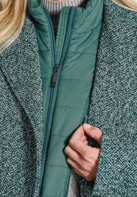 Eine Hand, die eine grüne gesteppte Jacke unter einem grünen gemusterten Mantel zippt, der über einem grauen Strickpullover getragen wird.