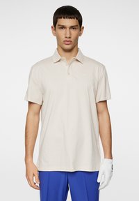 Kortärmad beige polo t-shirt gjord av bomull. Har en krage och en liten logotypdetalj på vänster bröst. Matchas med blå byxor.