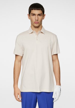 Short-sleeve beige polo shirt made of cotton. Features a collared neck and small logo detail on the left chest. Matched with blue trousers.