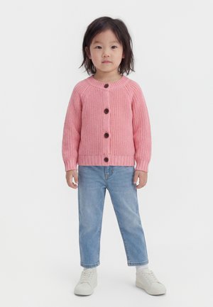 Νεαρό παιδί με σκούρα μαλλιά, φορώντας ροζ πλεκτή ζακέτα, ανοιχτόχρωμα μπλε τζιν και λευκά αθλητικά παπούτσια, στέκεται μπροστά σε απλό λευκό φόντο.