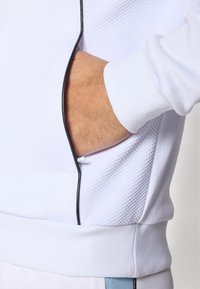 Top deportivo blanco con panel texturizado, cremallera negra contrastante y puños acanalados con acabado suave. Bolsillo accesible en el lateral.