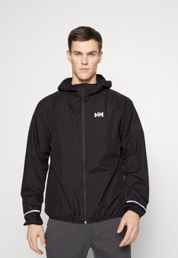 Schwarze wasserdichte Jacke mit Kapuze, ausgestattet mit einem Frontreißverschluss, elastischen Bündchen und einem reflektierenden Streifen an den Ärmeln. Logo auf der Brust.