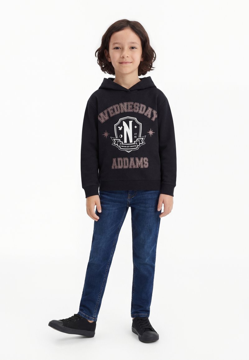 Παιδί με μαλλιά μέχρι τους ώμους, που φοράει μαύρο φούτερ Wednesday Addams, μπλε τζιν και μαύρα αθλητικά, στέκεται μπροστά σε λευκό φόντο.