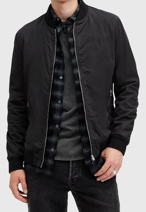 Mężczyzna w czarnej kurtce bomberce na czarno-białej koszuli w kratę i szarym t-shircie, ubrany w ciemne dżinsy, z ręką spoczywającą blisko kieszeni.