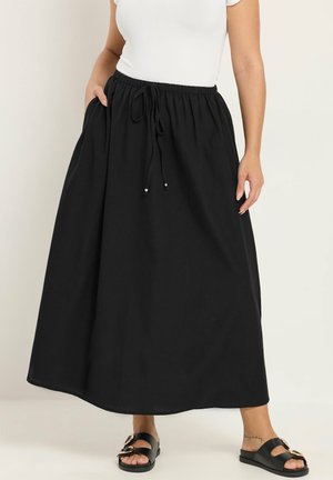 Vrouw draagt een lange zwarte rok met trekkoord en zijzakken, een wit kort mouwtopje en zwarte sandalen met dubbele gespen, staand op een witte vloer.