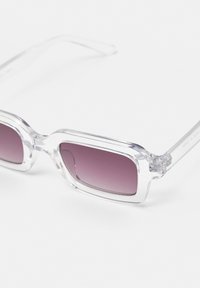 Ochelari de soare cu ramă transparentă și lentile violet degradé. Formă pătrată cu brațele groase. Designul notabil al articulației adaugă detaliu aspectului general.