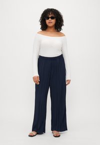 Navy pleated wide-leg pants with an elastic waistband, paired with a fitted white off-shoulder long-sleeve top and black sunglasses.