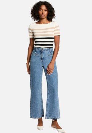 Haut en tricot à rayures beige, crème et noir ; jeans large taille haute bleu clair. Le mannequin porte des ballerines blanches à bout pointu. Cheveux bouclés.