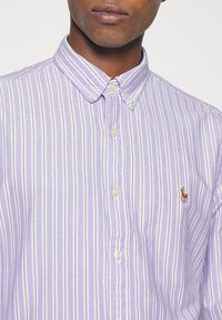 Chemise boutonée lilas clair avec des rayures blanches verticales, dotée d'un col classique, de boutons blancs et d'un petit logo brodé sur la poitrine.