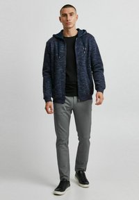 Hoodie zippé bleu marine avec cordons réglables, associé à un t-shirt noir et un pantalon gris ajusté. Des baskets noires décontractées complètent l'ensemble.