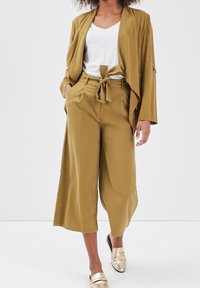 Sennepsgul blazer med brede ærmer, matchende højttalede culottes og en hvid T-shirt underneath. Guldsko uden hæl fuldender outfittet.