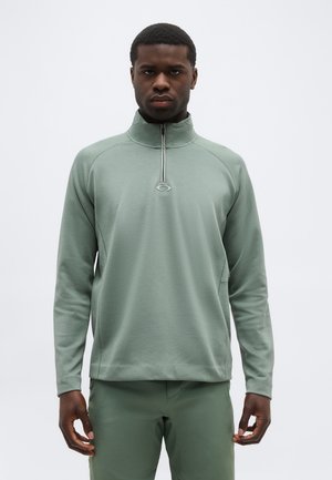 Mann trägt einen salbeigrünen Langarmpullover mit Viertelreißverschluss und passende Hose, steht vor einem schlichten, hellen Hintergrund.