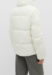 Vatteret jakke i blød hvid farve med quiltet design, høj krave og en glat, skinnende tekstur. Kombineret med grå bukser.