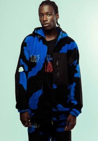 Chaqueta de forro polar con camuflaje azul y negro, cierre de cremallera, detalles bordados y capucha. El modelo lleva una camiseta gráfica negra debajo.