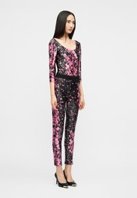 Γυναίκα που φοράει εφαρμοστό ολόσωμο φόρεμα με ροζ και μαύρο μοτίβο, με μανίκια μέχρι τον αγκώνα και μαύρες ψηλοτάκουνες γόβες, στέκεται σε απλό φόντο.