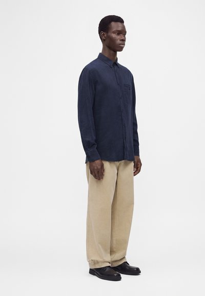 Man standing wearing a navy button-up shirt, beige wide-leg pants, and black shoes against a plain white background.