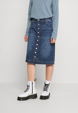 Personne portant une jupe en jean boutonnée jusqu'aux genoux, un haut bleu clair à manches longues, des bottes blanches à lacets et des chaussettes blanches, debout sur un fond uni.