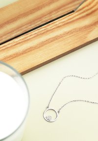 Collana in argento con pendente circolare decorato con un piccolo cuore, ornato di pietre scintillanti, posato su uno sfondo chiaro.