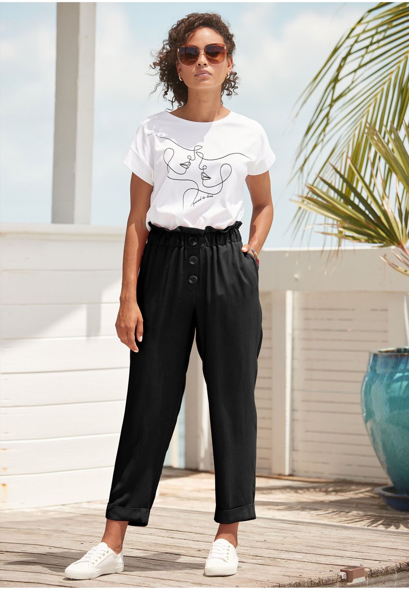 Weißes T-Shirt mit schwarzem Linienstil-Design, kombiniert mit schwarzen elastischen Hosen mit großen Knöpfen und hochgerollten Säumen. Weiße Sneakers runden den Look ab.