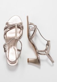 Une paire de sandales à talons ouverts en métal rose doré avec des fines brides, l'une montrée de dessus et l'autre de côté sur un fond blanc.