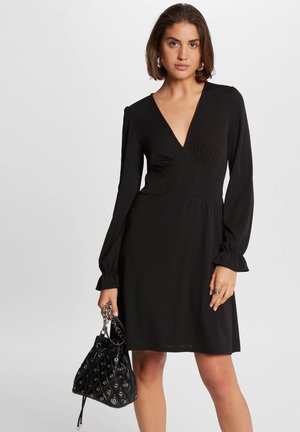 Robe noire à manches longues, décolleté en V profond et taille froncée. Assortie à un sac noir avec un motif matelassé et des accents en argent.