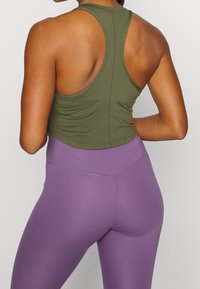 Žena v olivově zeleném tílku s racerback a vysokými fialovými legínami, zobrazená zezadu na neutrálním pozadí.
