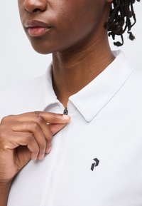 Κοντινό ενός ατόμου με σκούρο δέρμα που κλείνει ένα λευκό πουκάμισο με γιακά και μαύρο γεωμετρικό λογότυπο στο στήθος.