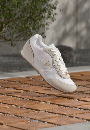Baskets blanches et beiges avec des lacets et un logo flottant au-dessus d'un sol en bois carrelé à l'extérieur.