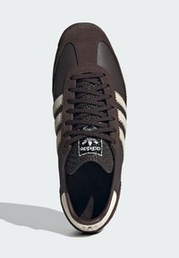 Tamno smeđi Adidas tenisice s kremastim prugama, teksturiranom jezičkom i crnim vezicama, prikazane odozgo na neutralnoj pozadini.