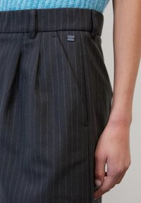 Tmavě pruhované kalhoty s přehybem vpředu, upraveným pasem a bočními kapsami. Materiál vypadá hladce s jemnou texturou.