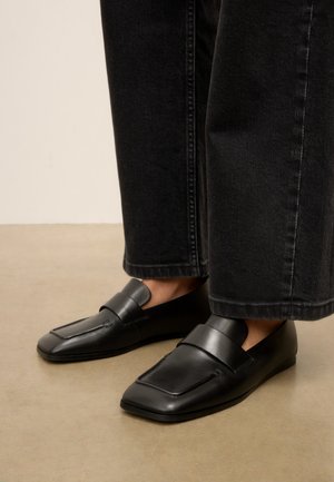 Zwarte leren slip-on loafers gedragen met zwarte recht gesneden jeans, staand op een beige vloer.