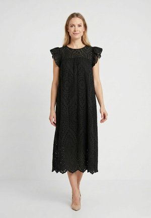 Femme portant une robe midi noire avec des détails en dentelle à œillets et des manches courtes à volants, associée à des chaussures à talons hauts beiges, se tenant devant un fond blanc.