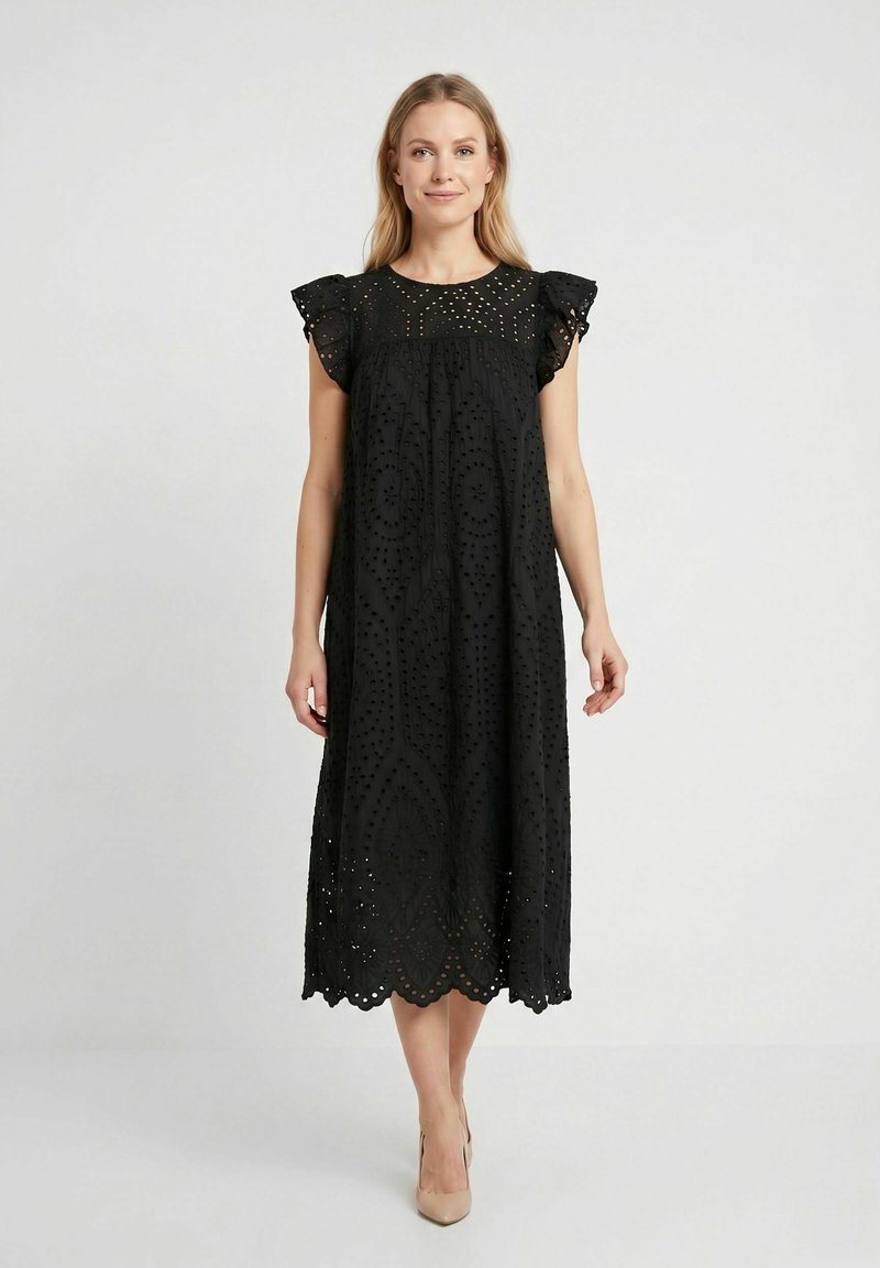 Femme portant une robe midi noire avec des détails en dentelle à œillets et des manches courtes à volants, associée à des chaussures à talons hauts beiges, se tenant devant un fond blanc.