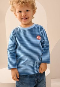 Kék és fehér csíkos hosszú ujjú póló kerek nyakkivágással, bal mellen hímezett vörös róka mintával. Farmernadrággal viselve.