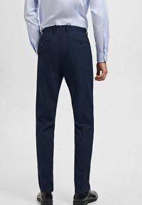 Pantalon ajusté bleu marine à coupe droite, avec poches arrière et texture subtile. Porté avec une chemise à col bleu ciel et des chaussures noires.