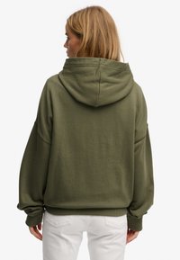 Felpa verde oliva con cappuccio grande, vestibilità oversize, polsini a costine e tessuto strutturato, abbinata a pantaloni bianchi.