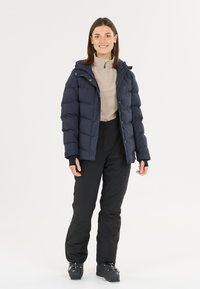 Marineblaue Daunenjacke mit Kapuze, ausgestattet mit einem gesteppten Design und Reißverschluss. Getragen über einem beigefarbenen Oberteil mit hohem Kragen und schwarzen Skihosen.