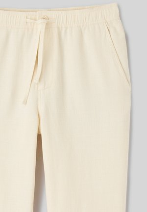 Pantalon texturé blanc cassé avec taille élastique, fermeture par bouton, cordon de serrage et poches latérales sur un fond gris clair.