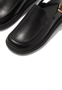Zwarte leren instappers met een gladde afwerking, ronde neus en minimale stiksels. Voorzien van een gouden hardwareaccent nabij de hak.