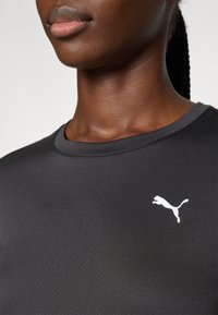 Svart atletisk skjorte med rund hals. Har en hvit Puma-logo på venstre bryst. Glatt, tettsittende stoff, designet for aktiv bruk.