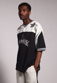 Jersey de gran tamaño en blanco y negro con acentos de estrellas, textura de malla y logo en contraste. Diseño de mangas cortas y escote en V con detalles a rayas.