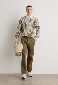 Knit sweater in earthy tones with abstract patterns, paired with olive green chinos. Model holds a beige jacket and wears white sneakers.