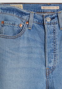 Jean en denim bleu avec poches avant, rivets en cuivre, passants de ceinture, fermeture par bouton métallique et une étiquette indiquant la taille "Ribcage Straight Ankle 27".