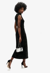 Czarna welurowa sukienka midi o dopasowanym kroju, wysokim dekolcie i rękawach typu cap, zestawiona z metaliczna srebrną torebką i czarnymi szpilkami.