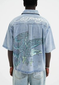 Lysblå denimskjorte med et stort broderet ørnerdesign i nuancer af grøn og blå, samt hvid "Ed Hardy" skrift på ryggen.